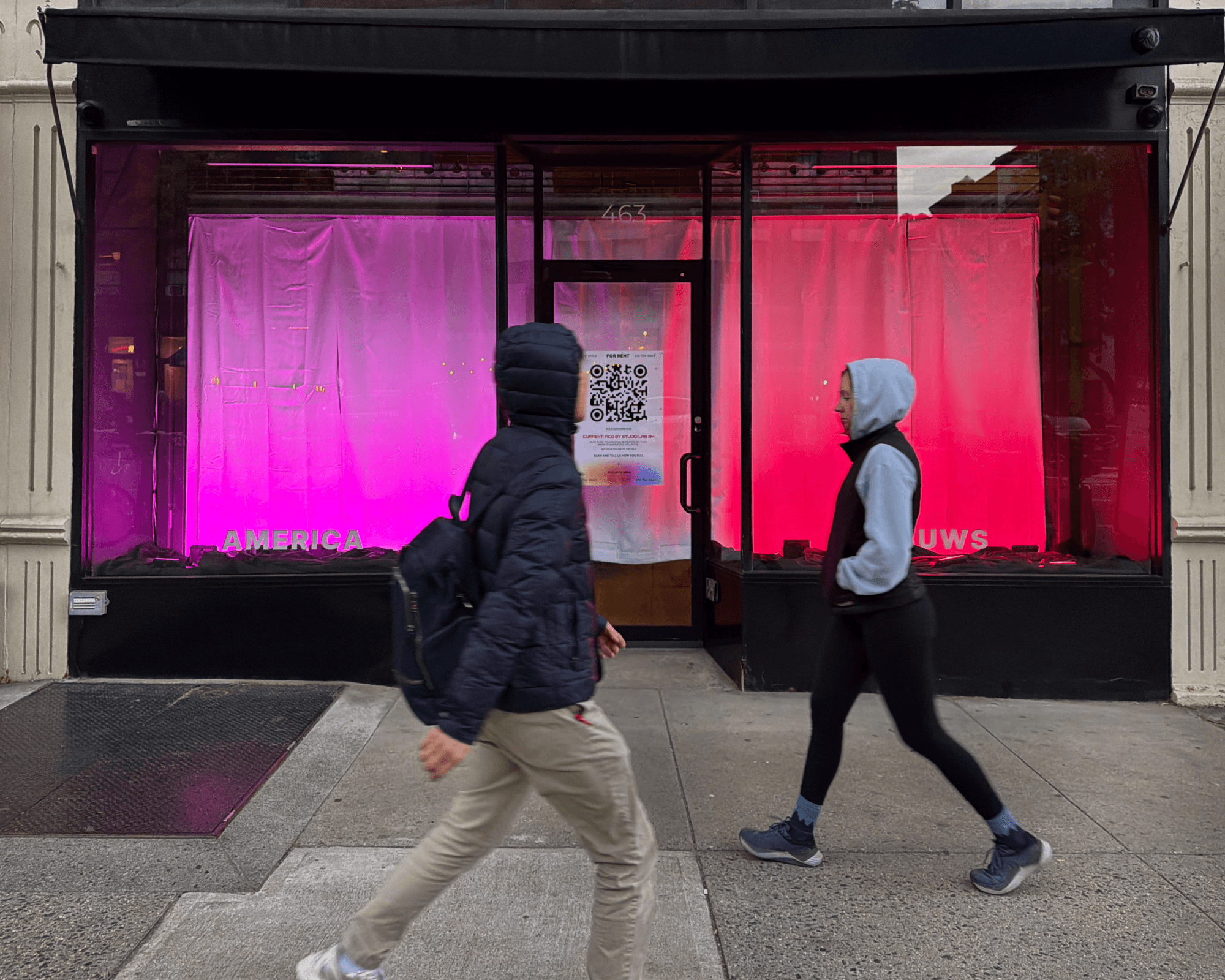 Two people pass the ACG storefront, with America lit violet and UWS lit red
